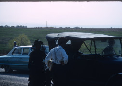 Actor Kyle Johnson (Newt Winger) and actress Hope Summers (Mrs. Kiner) stand left of black antique vehicle.