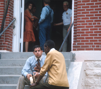 Actors talk on courthouse steps. Three actors stand inside doorway at top of stairs.