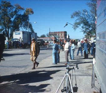 Actors Dana Elcar (Kirky) wearing a tan hat and Kyle Johnson (Newt Winger) in blue overalls, along with other actors and production crew in downtown Fort Scott, Kansas.