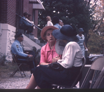 Actors and crew mingle outside of courthouse building. Two actresses talk while security officer sits next to courthouse stairs.