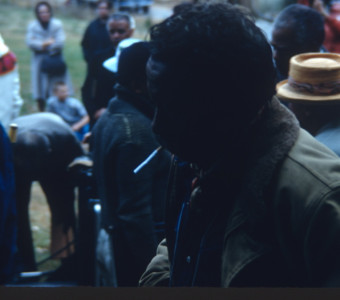 Director Gordon Parks, profile, smoking a cigarette. Actors and crew stand in background.