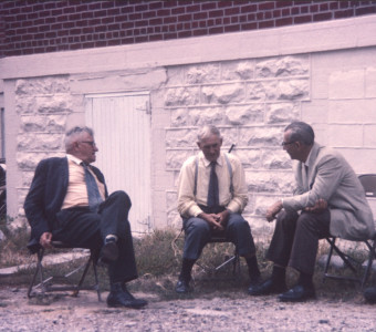 Actor Russell Thorson (Judge Cavanaugh) on left, and two other actors (likely playing jurors) sitting next to the courthouse in the trial scene.