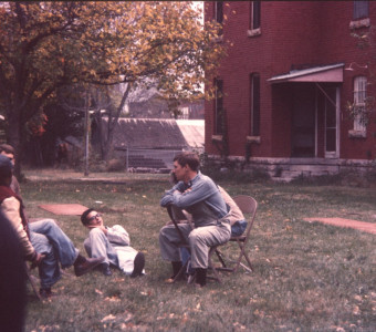 Five actors talking in the grass beside the courthouse building from trial scene.