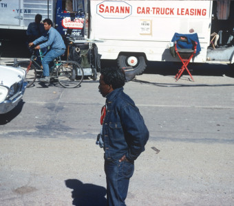Director Gordon Parks in front of production vehicles. Actor Stephen Perry (Jappy) rides a bicycle in the background.