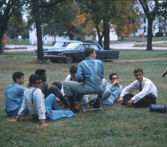 Seven actors sitting and relaxing in the grass while an actress standing is talking to them.