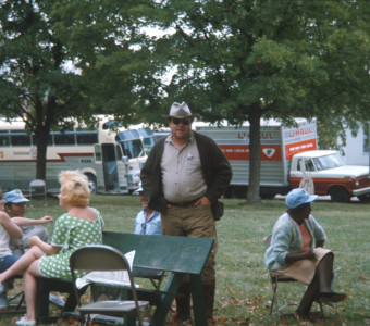Actor Dana Elcar (Kirky) stands in the middle of seated actresses in front of production vehicles.