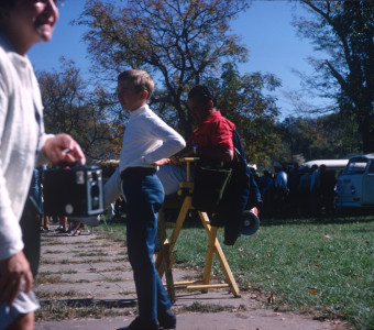 Production crew member, seated right, behind a young boy and woman with camera.