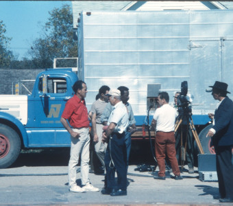 Production crew, including Director Gordon Parks, wearing blue jeans in the center of the photograph, and actor Malcolm Atterbury (Silas Newhall) standing beside a production truck.