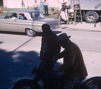 Director Gordon Parks stands alongside actor Dana Elcar (Kirky) who is seated on a motorcycle in front of production crew and equipment.