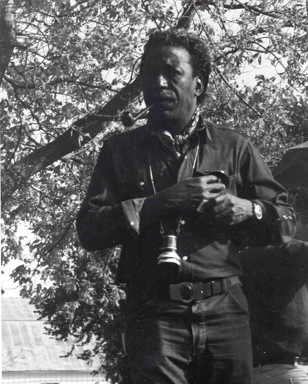Director Gordon Parks is smoking a pipe as he reaches for his shirt pocket. A camera lens hangs from his neck.