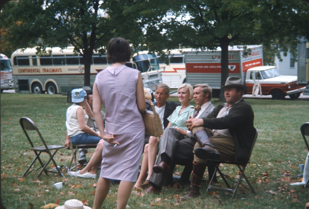 Actress with back to camera facing seated actors for The Learning Tree Actors, including Malcolm Atterbury (Silas Newhall), Don Dubbins (Harley Davis), and Dana Elcar (Kirky), sit in front of production vehicles.
