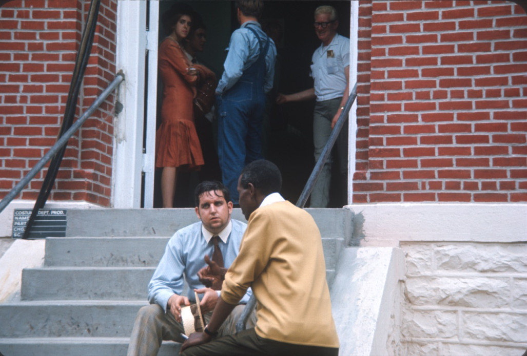 Actors talk on courthouse steps. Three actors stand inside doorway at top of stairs.