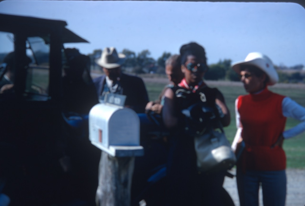 Actress Carol Lamond (Big Mabel), wearing round sunglasses, and other actors and crew prepare for a scene outside of Winger Home. The mailbox reads "Alex Mason."
