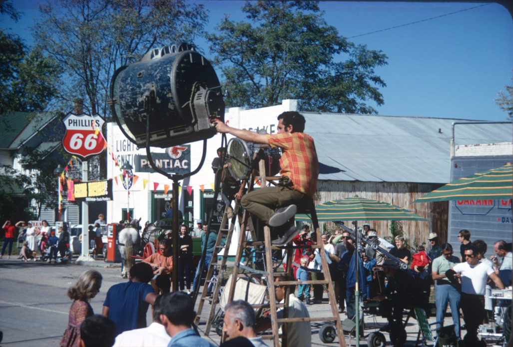 Actors and production crew filming in downtown Fort Scott, Kansas. One crew member is seated atop a tadder positioning a piece of lighting equipment.