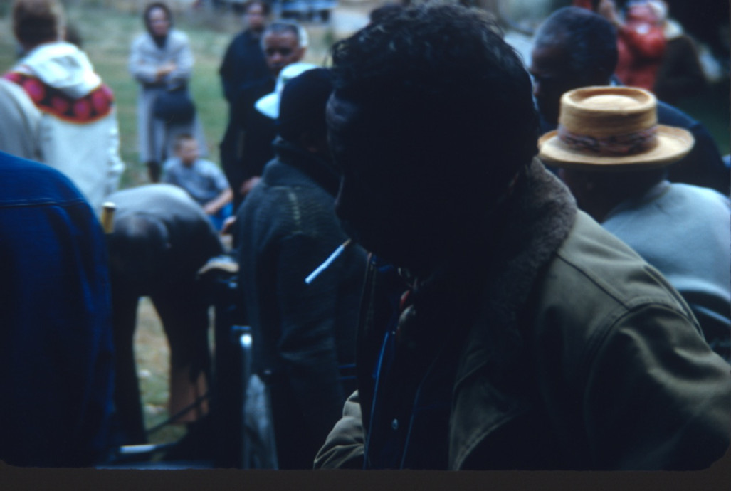 Director Gordon Parks, profile, smoking a cigarette. Actors and crew stand in background.