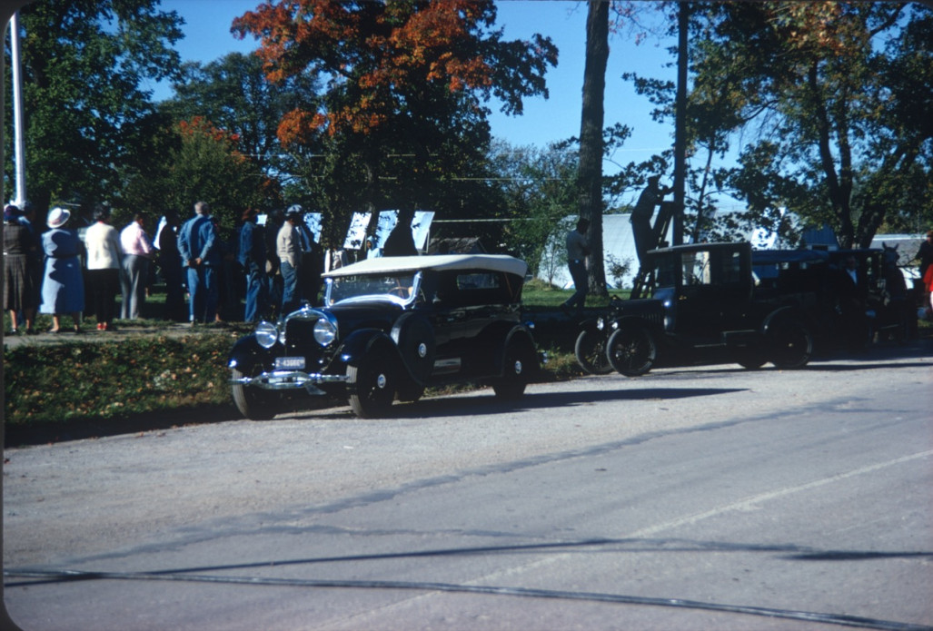 Antique cars parked along the street with actors in the background.