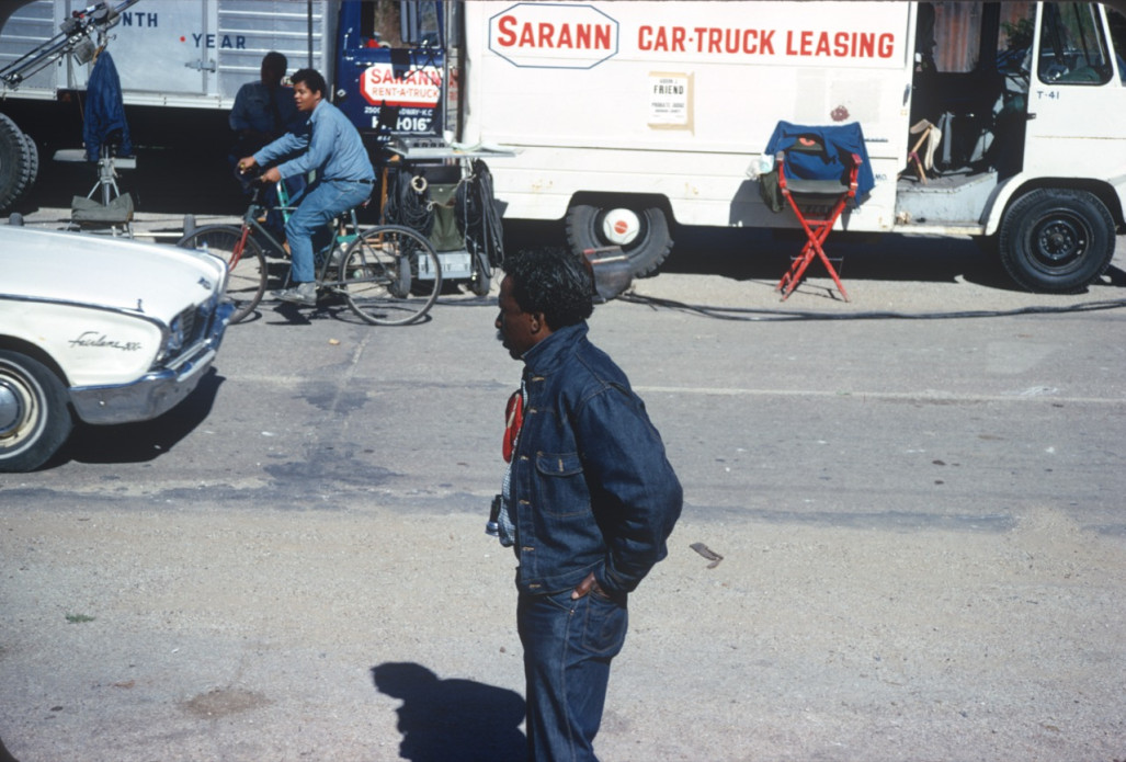 Director Gordon Parks in front of production vehicles. Actor Stephen Perry (Jappy) rides a bicycle in the background.