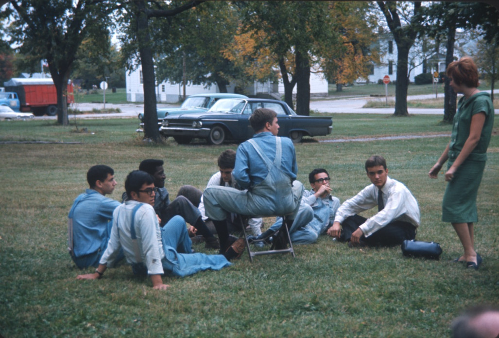Seven actors sitting and relaxing in the grass while an actress standing is talking to them.