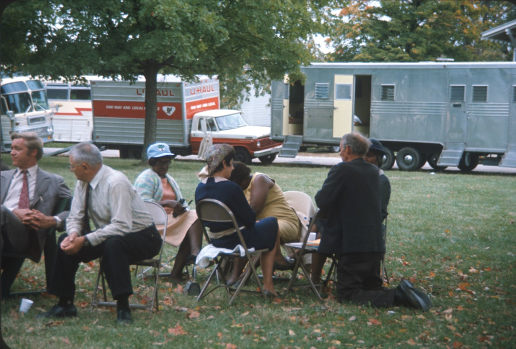Actors, including Estelle Evans (Sarah Winger), obscured by actor Malcolm Attebury (Silas Newhall) and Don Dubbins (Harley Davis), far left, seated in front of production vehicles.