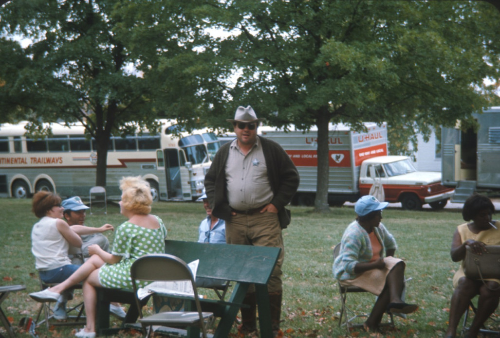 Actor Dana Elcar (Kirky) stands in the middle of seated actresses in front of production vehicles.