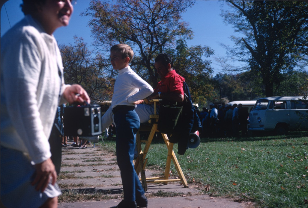 Production crew member, seated right, behind a young boy and woman with camera.