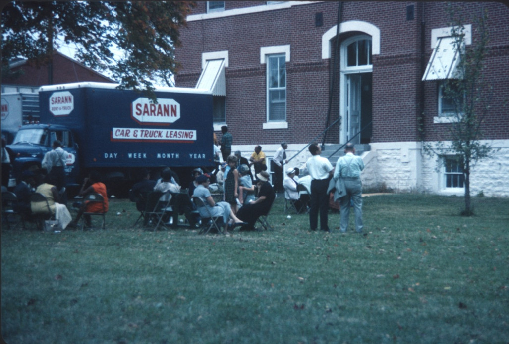 Learning Tree cast and crew sitting in groups outside of courthouse with production trucks Actors and crew members on a break outside of the courthouse used for the trial scene.