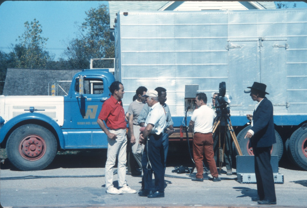 Production crew, including Director Gordon Parks, wearing blue jeans in the center of the photograph, and actor Malcolm Atterbury (Silas Newhall) standing beside a production truck.