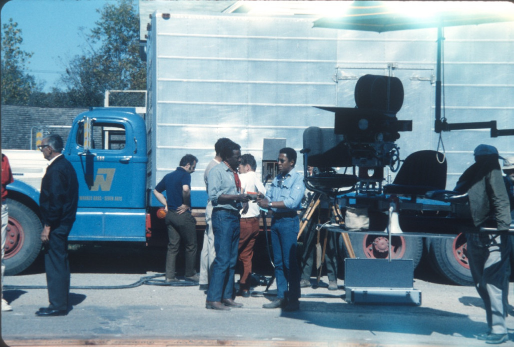 Director Gordon Parks, standing in the center of the photograph wearing a red ascot and blue jeans, speaks to his eldest son, Gordon Parks Junior, in front of production truck.