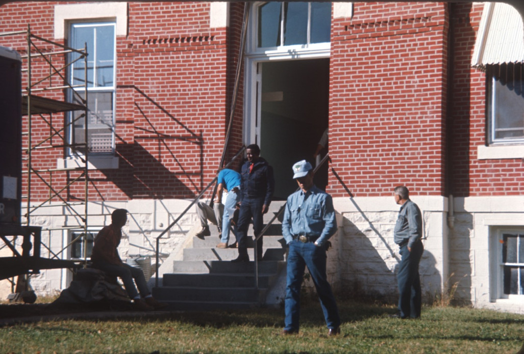 Director Gordon Parks walking down the steps of the courthouse building used for the trial scene with crew members milling about outside.